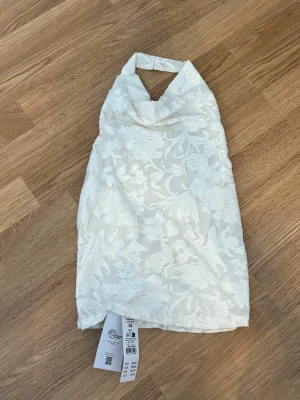 Vit halterneck topp med blommönster Gina - Snygg vit halterneck topp med diskret blommönster i struktur från ginatricot. Toppen har hög krage och är ärmlös, perfekt för sommarens varma dagar. Tillverkad i ett lätt och luftigt material som ger en elegant look.