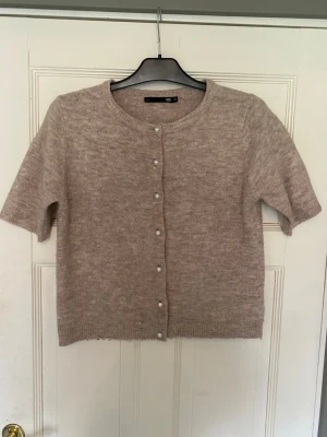 Beige kortärmad kofta med pärlknappar - Söt beige kofta från Vila med korta ärmar och rund halsringning. Koftan har fina pärlknappar framtill och är stickad i ett mjukt material som känns skönt mot huden. Perfekt att slänga över en topp för en chill look.