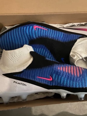 Nike Phantom 6 elite  - Säljer ett par Nike Phantom 6 elite fotbollsskor med högt skaft. Skorna har en snygg blå och svart ovandel med vita detaljer och rosa Nike-loggor. Ovandelen är stickad och ger en tajt passform, perfekt för snabba vändningar på planen.