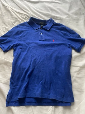Blå pikétröja från Polo Ralph Lauren - Klassisk blå pikétröja från Polo Ralph Lauren med korta ärmar och krage. Tröjan har två knappar framtill och den ikoniska röda logotypen broderad på bröstet. Tillverkad i mjuk bomull som känns skön mot huden. Perfekt till jeans eller shorts.