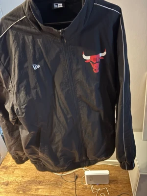 Chicago Bulls svart vindjacka New Era - Snygg svart vindjacka från New Era med Chicago Bulls-logga på bröstet och stort 'BULLS'-tryck på ryggen. Jackan har vita detaljer längs axlarna, dragkedja framtill och elastiska muddar. Perfekt för dig som gillar sportig streetwear och basketstil. Säljer för att jag har tappat mycket vikt och den är för stor helt enkelt