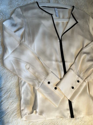 Blus med svarta detaljer från Mango - Stilren blus från Mango med svarta kontrastkanter längs knäppningen och halsringningen. Blusen har långa ärmar med svarta knappar. Tillverkad i ett glansigt, mjukt material som ger en elegant känsla. Använd 1 gång.