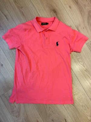 Ralph Lauren pikétröja - Säljer en klassisk rosa pikétröja från Ralph Lauren med mörkblå broderad logga på bröstet. Tröjan har korta ärmar, krage och knappar framtill. Perfekt för en avslappnad men ändå stilren look.