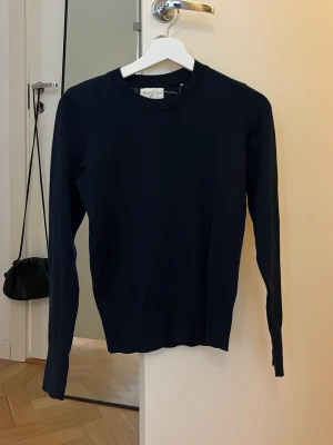 Mörkblå stickad tröja med gulddetaljer - Snygg mörkblå stickad tröja från Soft Goat med rund halsringning och långa ärmar. Tröjan har ribbade muddar och tre guldfärgade knappar vid ärmsluten som ger en exklusiv touch. Perfekt till jeans eller kjol för en stilren look.