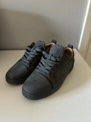 Gråa sneakers från Christian Louboutin - Snygga gråa sneakers från Christian Louboutin med klassisk röd sula och diskret logga undertill. Skorna har en låg profil, rund tå och är tillverkade i mocka med matchande grå skosnören. Insidan är fodrad i beige läder för extra komfort.