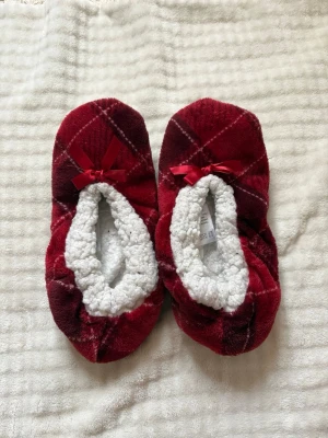 Rödrutiga tofflor med rosett - Mysiga tofflor i röd- och vinrutig plysch med små rosetter framtill. Insidan är fodrad med mjuk, vit teddy som håller fötterna varma. Yttersulan är i beige mockaimitation. Perfekta för att chilla hemma under kalla dagar.