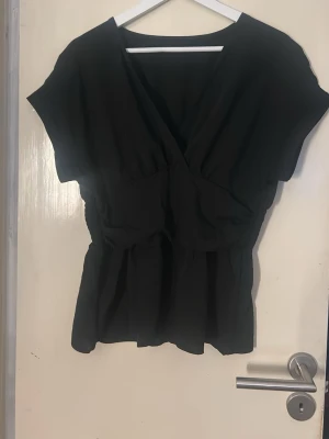 Svart v-ringad blus med kort ärm - Stilren svart blus med v-ringning och markerad midja. Blusen har korta ärmar och ett lätt, mjukt tyg som ger ett snyggt fall. Perfekt att matcha med både jeans och kjol för en trendig look.