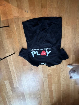 Svart Comme des Garçons PLAY t-shirt - Snygg svart t-shirt från Comme des Garçons PLAY med det ikoniska röda hjärtat och vit text på bröstet. Klassisk passform och rund hals. Perfekt för dig som gillar streetwear och vill ha något stilrent men ändå lekfullt.
