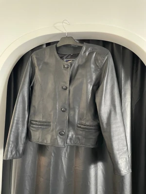 Svart skinnjacka utan krage - Stilren svart skinnjacka med rund halsringning och stora knappar framtill. Jackan har raka fickor och en kortare, boxig passform. Perfekt för dig som gillar minimalistisk och tidlös stil.