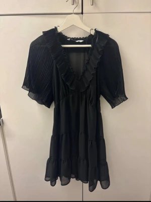 Svart volangklänning med plisserade ärmar - Snygg svart kortklänning med volangdetaljer och plisserade, luftiga kortärmar. Klänningen har v-ringning med bred volangkant och flera lager i kjolen för extra volym. Perfekt för dig som gillar en romantisk och trendig stil.