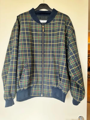 Rutigt bomberjacka från Lou Atkins - Bomberjacka med rutigt mönster i blått, gult, grönt och rött. Jackan har dragkedja framtill, två fickor. Klassisk bombermodell med avslappnad passform och mjukt material. Perfekt för dig som gillar retrostil. Köpt på secondhand. Lätt att ha på sig och luftig. 