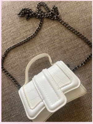 Vit miniaxelväska med kedja - 💞Säljer en liten vit axelväska i croco-mönstrat konstläder med silverfärgad kedjerem. Väskan har ett top handtag och en unik design med dubbla lockdetaljer framtill. Perfekt för dig som gillar trendiga accessoarer och vill sticka ut.💕