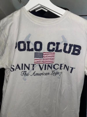 Vit Polo Club Saint Vincent t-shirt - Vit t-shirt från Polo Club Saint Vincent med tryck framtill i blått och rött samt amerikansk flagga. Klassisk rund halsringning och korta ärmar. Tillverkad i mjuk bomull, perfekt för en avslappnad stil.