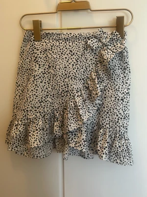 Två volangkjolar - Säljer två korta kjolar med volang och rosett. Ena kjolen är vit med svart prickigt mönster, den andra är blå med små vita blommor. Båda har omlott-look och volangkant nertill. Perfekta för sommarens alla tillfällen!