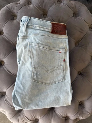 Ljusblå slitna jeans från Replay - Säljer ett par ljusblå jeans från Replay med slitningar och distressed detaljer. Klassisk femficksmodell med läderpatch bak i midjan och röd Replay-logga på bakfickan. Jeansen har en avslappnad passform och är tillverkade i bomull.