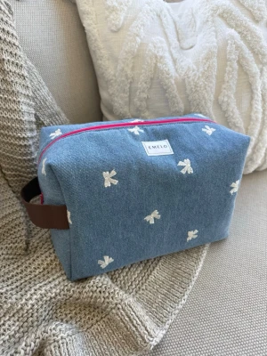Blå jeans necessär med broderade rosetter - Söt necessär från Emelo i blått jeanstyg med vita broderade rosetter. Väskan har en rosa dragkedja och ett brunt handtag på sidan. Perfekt för smink eller småprylar. Tygmaterialet ger en avslappnad och trendig känsla. Storlek large, mått ca 27x17x12cm