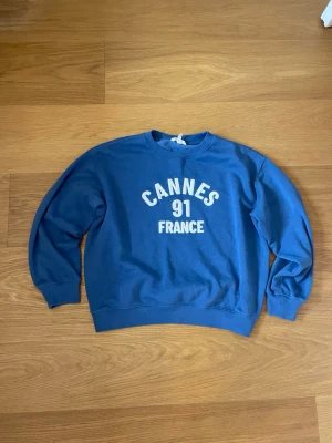 Blå sweatshirt med Cannes-tryck - Blå sweatshirt med vitt tryck där det står 'Cannes 91 France' på bröstet. Tröjan har rund halsringning, ribbade muddar vid ärmslut och nederkant. Skön