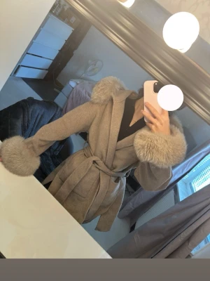 Beige kappa  - Snygg beige kappa med stora räv pälskrage på huva och ärmslut. Kappan har knytskärp i midjan och en mjuk, figurnära passform. Perfekt för dig som vill ha en trendig och mysig look under kalla dagar. Knappast använd köpt från Furstockholm stl xs-m