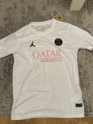 Vit PSG matchtröja Jordan x Nike - Säljer en vit PSG matchtröja med rosa detaljer och Qatar Airways-tryck på bröstet. Tröjan har Paris Saint-Germain klubbmärke och Jordan-logga framtill. Tillverkad i lätt och ventilerande material, perfekt för fotboll eller träning.