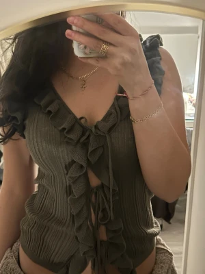 Olivgrön ribbad topp med volang - Trendig olivgrön ribbad topp med volangdetaljer längs v-ringningen och framtill. Toppen har knyt framtill och är ärmlös, perfekt för en snygg och avslappnad stil.