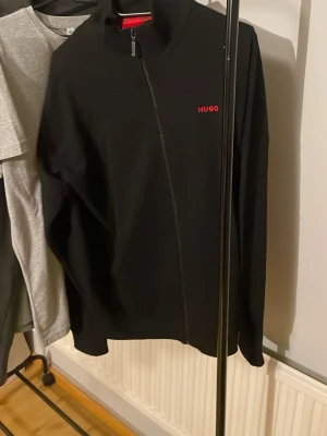 Svart stickad tröja från HUGO - Snygg svart stickad tröja från HUGO med dragkedja och röd logga broderad på bröstet. Tröjan har hög krage och långa ärmar, perfekt för en clean och stilren look. Materialet känns mjukt och följsamt.