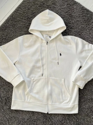 Vit hoodie från Polo Ralph Lauren - Säljer en clean vit hoodie från Polo Ralph Lauren med dragkedja och klassisk svart logga broderad på bröstet. Hoodien har fickor framtill, justerbar huva med snören och är gjord i mjukt bomullsmaterial. Perfekt för en chill och stilren look.