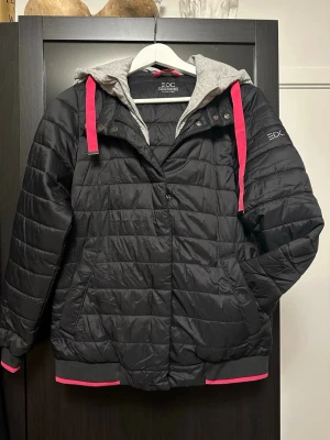 Svart pufferjacka från EDC med huva (ESPRIT) str. M - Snygg svart pufferjacka från EDC med grå huva och rosa detaljer på snören och muddar. Jackan har quiltad design, dragkedja och knappar framtill samt två sidofickor. Innerfodret är grått och mjukt, vilket ger extra komfort och stil. Innefodret samt luvan går att ta bort. 