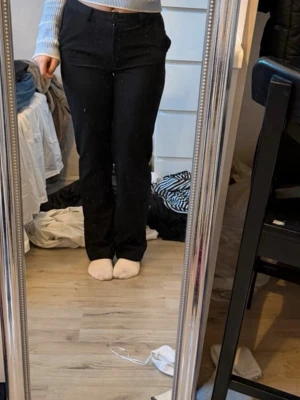Svarta kostymbyxor med rak passform - Säljer ett par klassiska svarta kostymbyxor med hög midja och rak passform. Byxorna har hällor för skärp och stängs med knapp och dragkedja. Perfekta till en snygg topp eller skjorta. Materialet känns mjukt och följsamt.