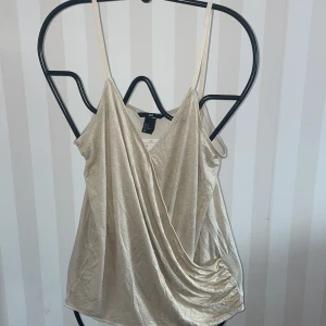 Beige glittrig linnetopp från H&M Ny - Säljer en snygg beige glittrig linnetopp från H&M. Toppen har smala axelband och en omlott-design framtill som ger en elegant touch. Perfekt för att lysa upp vilken outfit som helst! ✨