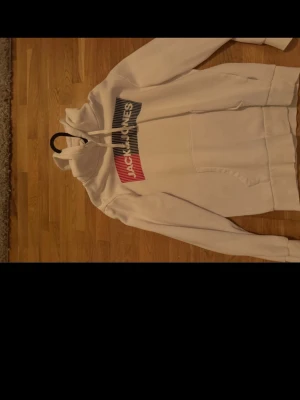 Vit hoodie från Jack & Jones - Snygg vit hoodie från Jack & Jones med stor magficka och dragsko i huvan. Tryck på bröstet i blått, vitt och rött med loggan. Perfekt för en avslappnad och trendig stil. Mjuk insida och klassisk passform.