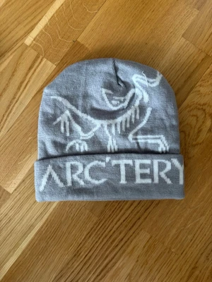 Grå mössa från Arc'teryx - Snygg grå mössa från Arc'teryx med vit logga och grafik av fossil på framsidan. Mössan har uppvikt kant och är tillverkad i mjukt stickat material som passar perfekt för kyliga dagar.