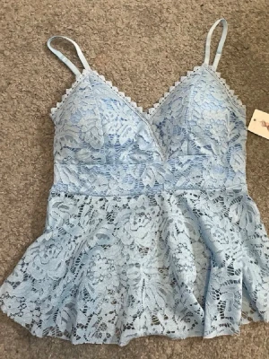 Blått linne - Söt ljusblå topp i spets med smala axelband och v-ringad hals. Blommigt spetsmönster över hela plagget och lätt utsvängd nederdel. Perfekt för dig som gillar romantisk och feminin stil.