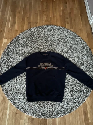 Mörkblå sweatshirt från Morris - Snygg mörkblå sweatshirt från Morris med broderad text 'Morris Sporting Club' och dekorativa linjer över bröstet. Klassisk rund halsringning och ribbade muddar vid ärmslut och nederkant. Perfekt för en avslappnad och stilren look.
