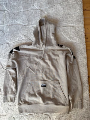 Beige hoodie från Adidas med tryck - Snygg beige hoodie från Adidas med stor svart logga över ryggen och svarta detaljer på axlarna. Klassisk känguruficka framtill och justerbar huva med dragsko. Tillverkad i mjuk bomullsblandning, perfekt för en avslappnad streetwear-look.