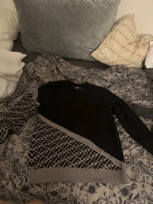Svart och grå stickad Fendi-tröja - Säljer en stickad tröja från Fendi i svart och grått med det klassiska Fendi-mönstret på ena ärmen och nedre delen av tröjan. Tröjan har rund halsringning och ribbade muddar. Perfekt för dig som gillar exklusiva märken och unik design.