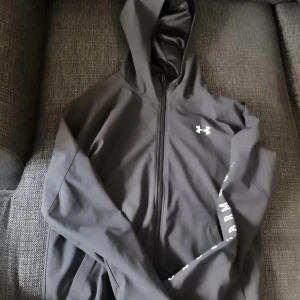 Mörkblå vindjacka från Under Armour XS - Svart vindjacka från Under Armour med huva och hel dragkedja. Jackan har logga på bröstet och stort tryck längs ena ärmen. Lätt material och loose fit, perfekt för aktiva dagar. Snygg och sportig stil som funkar året runt.