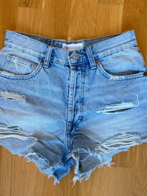 Blåa high waist jeansshorts från Zara - Säljer ett par ljusblå denimshorts från Zara med hög midja och slitna detaljer. Shortsen har fransiga benslut, klassiska fem fickor och knappgylf. Perfekta för varma dagar och ger en avslappnad look.