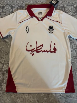 Vit fotbollströja Palestina - Säljer en vit fotbollströja från paliroots med vinröda detaljer och texten 'Palestina' på bröstet. Tröjan har klubbmärke och karta broderat på framsidan samt klassisk krage. Tillverkad i lätt och ventilerande material, perfekt för träning eller match.