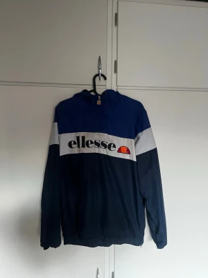 Blå och vit vindjacka från Ellesse - Snygg vindjacka från Ellesse i blått och vitt med stor logga framtill. Jackan har huva, dragkedja och elastiska muddar. Perfekt för dig som gillar sportig stil och vill ha något lätt att slänga på sig. Materialet känns tunt och smidigt.