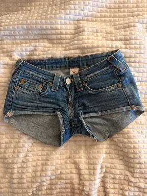 True religion jeansshorts (lowwaist) - Säljer ett par väldigt snygga jeansshorts från True Religion i storlek 26.  Shortsen är egensydda och omgjorda, vilket gör dem helt unika. De är lågmidjade och korta med en riktigt snygg passform.  Klassisk True Religion denim med deras ikoniska bakfickor och kontrastsömmar som ger en tydlig Y2K/vintage känsla. De har slitna och råa detaljer som ger en cool och edgy look.  Sjukt snygga, men tyvärr för små för mig. (Mått kan skickas vid intresse)  • Märke: True Religion • Egensydda/omgjorda 