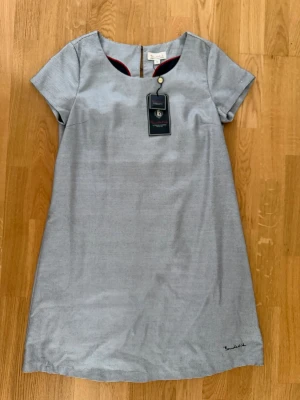 Grå kortklänning Bondelid - Stilren grå kortklänning från Bondelid med diskret rutigt mönster och rund halsringning med knappdetalj. Klänningen har korta ärmar och är tillverkad i ett mjukt bomullsmaterial. Perfekt för en avslappnad men snygg look.