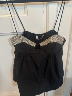 Svart topp med strassdetaljer Zara - Trendig svart topp från Zara med tunna axelband och glittrande strassdetaljer längs överdelen. Toppen har en lätt puffig passform och en liten cut-out framtill. Perfekt för dig som vill sticka ut med en elegant touch.