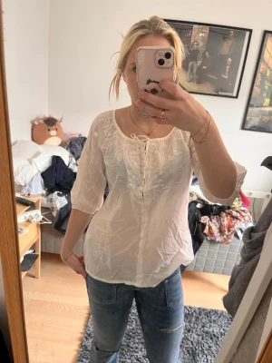 Vit blus med knytning fram - Säljer en luftig vit blus med trekvartsärm och dekorativ knytning vid halsen. Blusen har broderade detaljer framtill och är tillverkad i ett tunt, lätt material som ger en avslappnad look. Perfekt att matcha med jeans eller kjol.