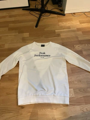 Vit sweatshirt Peak Performance - Snygg vit sweatshirt från Peak Performance med svart broderad logga och text 'Est. 1986' på bröstet. Klassisk rund halsringning och ribbade muddar vid ärmslut och nederkant. Perfekt för en avslappnad och stilren look. Den har två fläckar , men inget som syns och inget som jag försökt att ta bort så inte omöjligt att dom går bort. 
