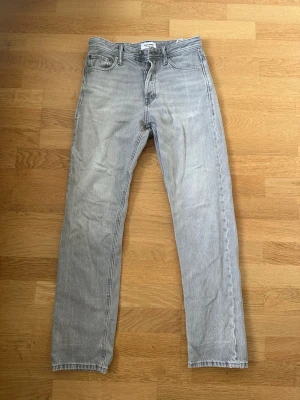 Ljusgrå raka jeans från Jack and Jones - Säljer ett par ljusgrå jeans från Jack And Jones med rak passform och klassisk femficksdesign. Jeansen har hög midja, gylf med knappstängning och är tillverkade i slitstark denim. Perfekta för en clean och avslappnad stil.