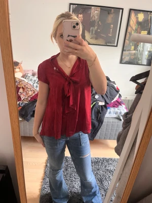 Vinröd blus med knyt framtill - Snygg vinröd blus med korta ärmar och knytband vid halsen. Blusen har en lös passform och ett mjukt, lätt tyg som faller fint. Perfekt för att lyfta en enkel outfit med lite färg och stil.