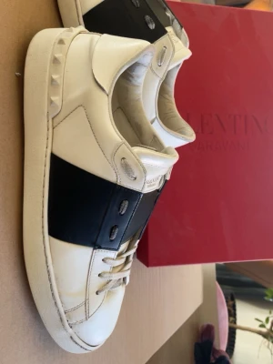 Valentino Garavani vita sneakers - Säljer ett par vita sneakers från Valentino Garavani med bred svart läderdetalj över mitten och nitar längs hälen. Det följer även med kartong, extra snören och 2 dustbags