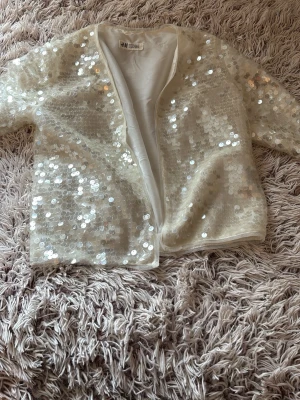 Vit paljettkofta från H&M - Säljer en glittrig vit kofta från H&M täckt med stora paljetter. Koftan har öppen front, trekvartsärmar och är fodrad på insidan för extra komfort. Perfekt för dig som vill sticka ut och addera lite extra bling till din outfit. Jätte söt kofta . Strolek 134-140 men passar även för någon med storlek xxs 