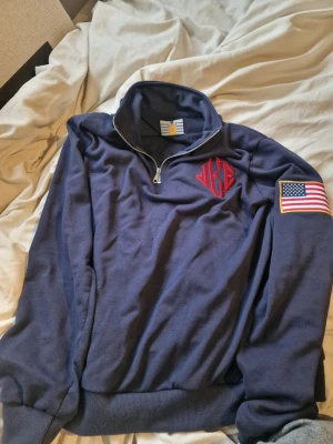 Jeffrey quarter zip - Snygg marinblå stickad tröja med halv dragkedja och hög krage. Tröjan har ett rött broderat monogram på bröstet och en amerikansk flagga på ärmen. Ribbstickade muddar vid ärmslut och nederkant. Perfekt för dig som gillar klassisk college-stil.