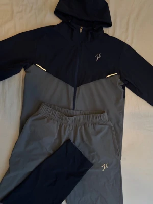 Mörkblå och grå träningsset från jeu d'ombres  - Snyggt träningsset från  jeu d'ombres i mörkblått och grått. Setet består av en zip-hoodie med huva och matchande byxor, båda med vita detaljer och logga på bröst och ben. Perfekt för träning eller chill. Materialet känns lätt och smidigt.